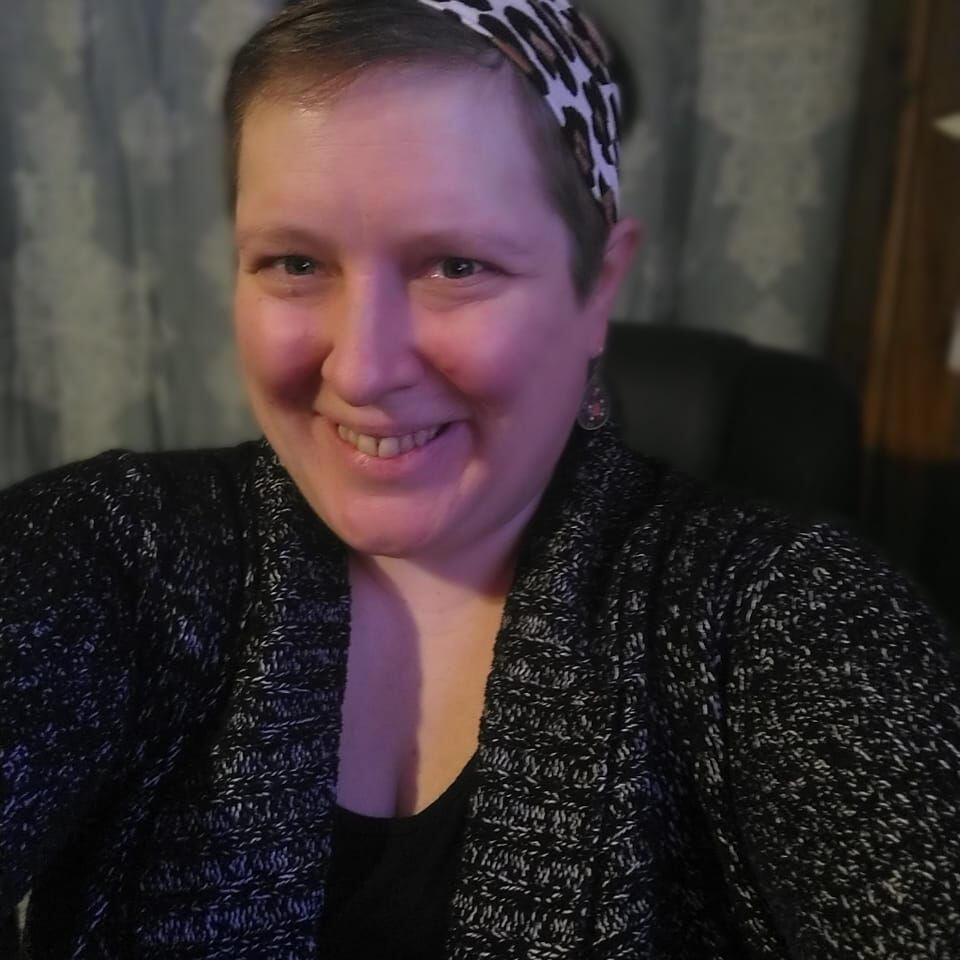 Woman in green hat, red shirt and black pants in front of mountain range with arms open